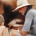 Homem idoso e criança ordenham uma vaca ao ar livre em uma propriedade rural brasileira.